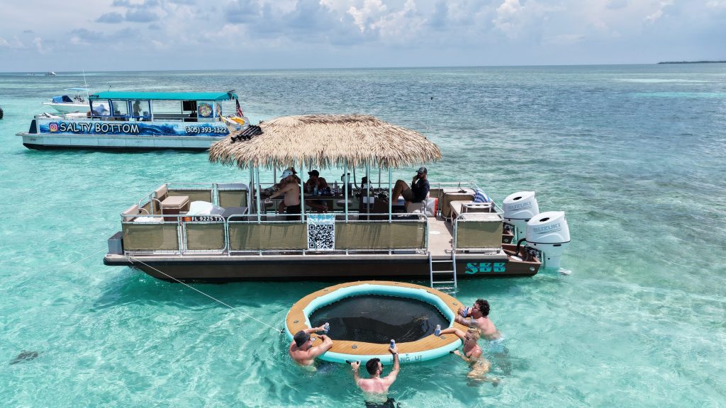 Aerial view of the tiki boat anchored in turquoise shallows, with guests on a floating trampoline and the party boat behind